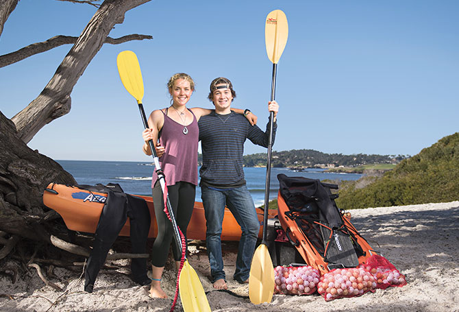 Two kids stand and smile next to their kayak and several bags of golf balls.