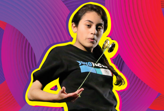 A young woman leaning back slightly while swinging a gold yo-yo upwards, against a pink background