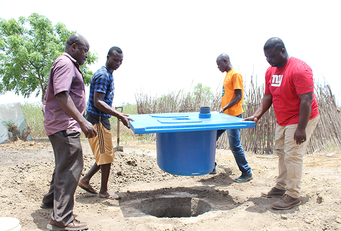 Four people carrying a blue toilet system into a hole